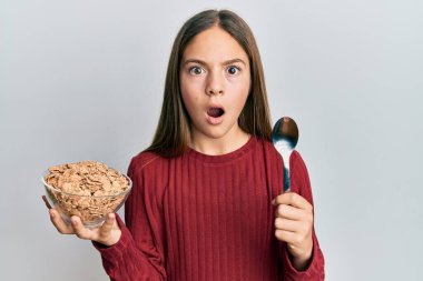 Güzel esmer küçük kız elinde kaşıkla sağlıklı tahıl gevrekleri yiyor. Şaşkınlık, şaşkınlık, korku ve heyecan dolu bir yüz ifadesi var.. 