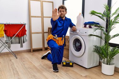 Genç İspanyol adam çamaşır makinesinde çalışıyor, eli açık, dur işareti yapıyor, ciddi ve kendinden emin bir ifadeyle, savunma hareketi yapıyor. 