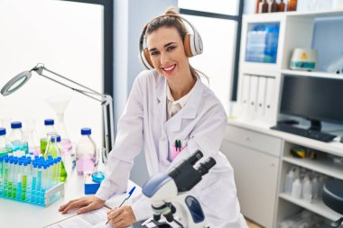 Bilim adamı üniforması giyen genç bir kadın laboratuarda müzik yazıları dinliyor.