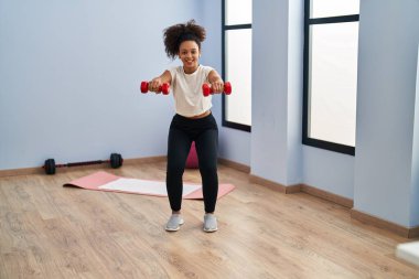 Genç Afrikalı Amerikalı kadın gülümsüyor spor merkezinde halterleri kullanarak kendine güven eğitimi alıyor