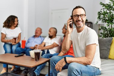Kanepede oturan bir grup orta yaşlı arkadaş konuşuyor. Gülümseyen mutlu bir adam evde akıllı telefondan konuşuyor..