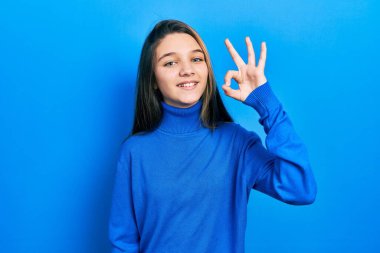 Balıkçı yaka kazak giyen genç esmer kız gülümsüyor ve el ve parmaklarıyla 