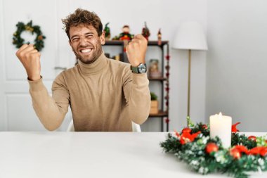 Noel süslemeleriyle masada oturan sakallı yakışıklı genç adam kolları açık ve gözleri kapalı bir şekilde başarının şaşkınlığını ve şaşkınlığını kutluyor. Kazanan konsept. 