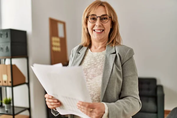 Middle age businesswoman smiling happy holding paperwork standing at the office.