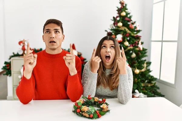 Young Hispanic Couple Sitting Table Christmas Amazed Surprised Looking Pointing — Stockfoto