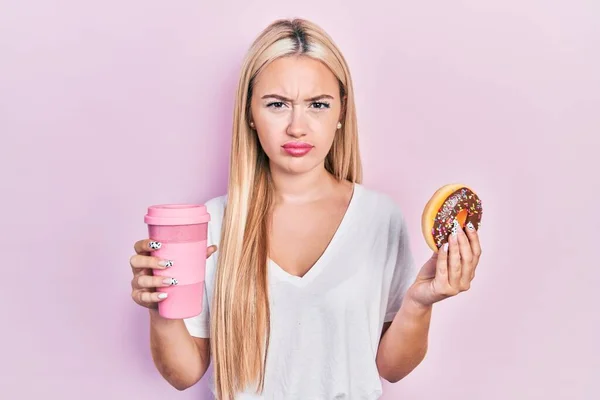 年轻的金发姑娘吃着甜甜圈 喝着怀疑和紧张的咖啡 因为问题皱着眉头 消极的人 — 图库照片