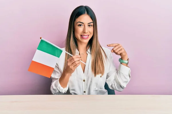 Jovem Hispânica Segurando Bandeira Irlandesa Sentada Mesa Sorrindo Feliz Apontando — Fotografia de Stock