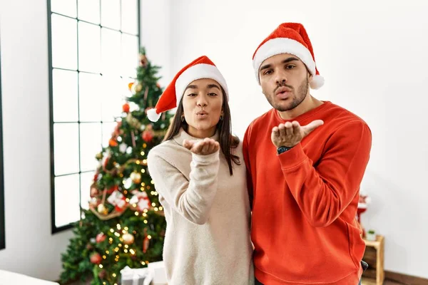 カメラを見ているクリスマスツリーのそばに立つ若いヒスパニック系のカップルは 愛とセクシーさを空中で手でキスを吹いています 愛の表現 — ストック写真