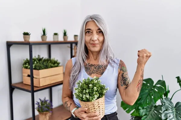 家庭で緑の植物の鍋を保持している中年の白髪の女性は非常に満足し 腕を上げて勝者のジェスチャーを行う興奮 笑顔と成功のために叫んで お祝いのコンセプト — ストック写真