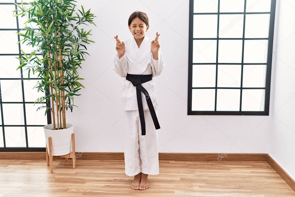 Young hispanic girl wearing karate kimono and black belt gesturing finger crossed smiling with hope and eyes closed. luck and superstitious concept. 