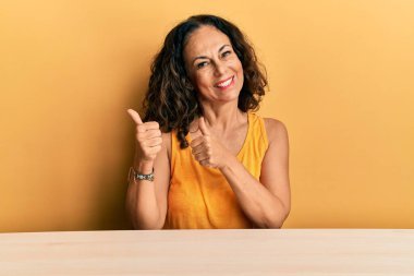 Güzel orta yaşlı kadın, gündelik giysiler içinde masanın üzerine oturmuş, arkasını işaret ediyor, el ve baş parmakları havada, kendinden emin bir şekilde gülümsüyor. 