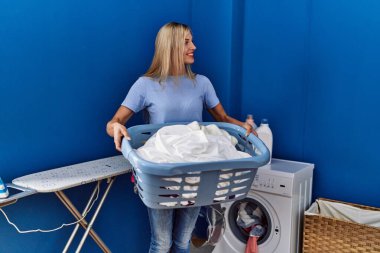 Çamaşır odasında kendine güvenen, elinde sepet tutan, gülümseyen genç sarışın kadın.