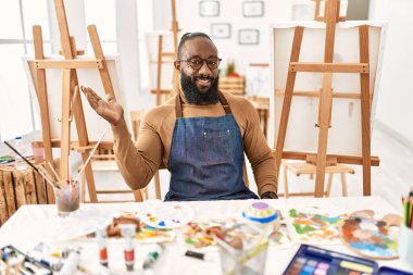 African american artist man at art studio smiling cheerful presenting and pointing with palm of hand looking at the camera. 