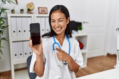 Klinikte elinde akıllı telefon tutan doktor üniforması giyen genç Latin kadın.
