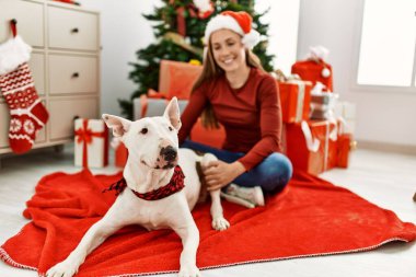 Beyaz bir kadın gülerek evde köpekle oturuyor. Noel ağacı yanında.