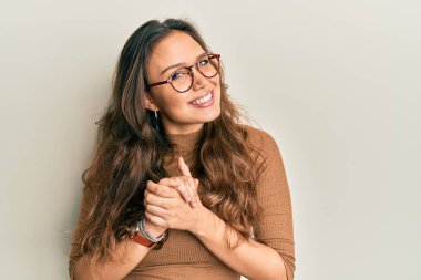 Latin asıllı genç bir kız, günlük kıyafetler ve gözlük takıyor ve ellerini kavuşturup gülümsüyor. Rahatlamış ve neşeli. Başarı ve iyimserlik 