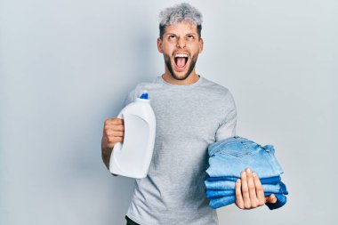Modern boyalı saçları olan genç İspanyol bir adam çamaşır için kot pantolon tutuyor ve çamaşır deterjanı şişesi kızgın ve kızgın bağırıyor sinirli ve öfkeli, öfkeyle bağırıyor.. 