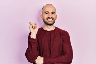 Yüzünde kocaman bir gülümseme olan, sıradan kıyafetler giyen genç kel bir adam, kameraya el ve parmak ile bakıyor.. 