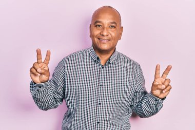 Orta yaşlı, sıradan kıyafetler giyen, gülümseyen, parmaklarını zafer işareti yaparken gösteren Latin bir adam. İki numara.. 