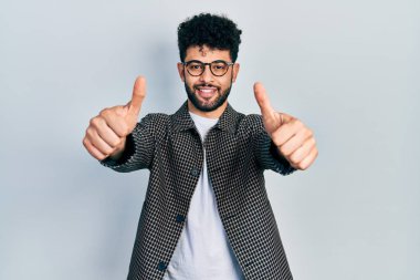 Sakallı, gözlüklü genç bir Arap. Elleriyle olumlu bir jest yapıyor. Gülümsüyor ve başarı için seviniyor. kazanan jest. 