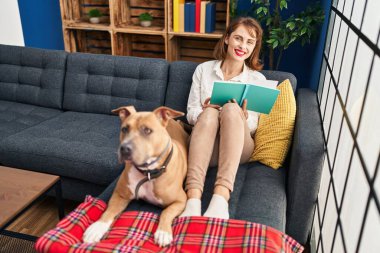 Genç beyaz kadın kitap okuyor. Kanepede köpeğiyle oturuyor.