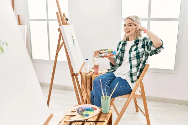 若いアーティストの女性は 顔の上に指で平和のシンボルを行うアートスタジオでキャンバス上の絵 勝利を示す明るい笑顔 — ストック写真