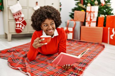 Genç Afrikalı Amerikalı kadın kahve içiyor ve kitap okuyor. Evdeki Noel ağacının yanında yatıyor.