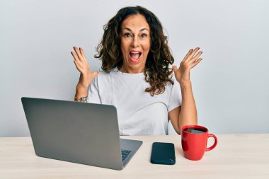 Ofiste çalışan orta yaşlı güzel bir kadın bilgisayarını kullanarak zaferi mutlu bir gülümsemeyle ve ellerini kaldırarak kutluyor. 