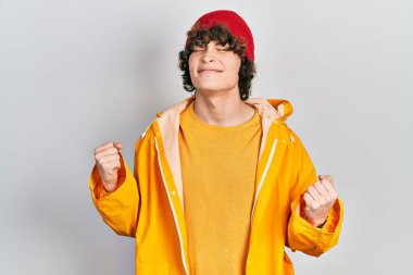 Sarı yağmurluk giyen yakışıklı genç adam çok mutlu ve heyecanlı kolları havada, gülümseyen ve başarı için bağıran kazanan bir jest yapıyor. kutlama konsepti. 