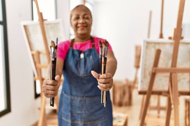 Kıdemli Afro-Amerikan kadın resim stüdyosunda elinde fırçayla gülümsüyor.