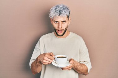 Modern boyalı saçları olan genç İspanyol bir adam şok olmuş bir fincan kahve içiyor, şüpheci ve alaycı görünüyor, ağzı açık kalmış. 