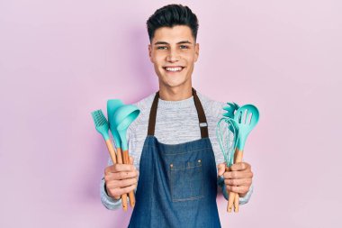 Profesyonel fırıncı önlüğü giyen genç İspanyol adam elinde yemek aletleri tutarken yüzünde mutlu ve havalı bir gülümsemeyle gülümsüyor. diş gösterme. 