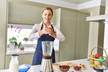 Genç sarışın kadın mutfaktaki blendere muz dökerek gülümsüyor.