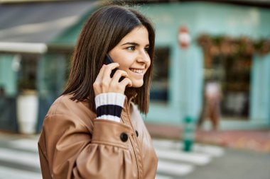 Genç, güzel, esmer bir kadın dışarıda mutlu bir şekilde gülümsüyor telefonda konuşuyor.