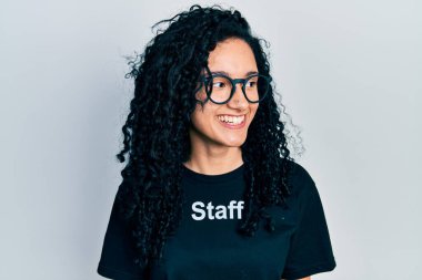 Kıvırcık saçlı, personel tişörtlü, yüzünde gülümsemeyle yan yana bakan İspanyol bir kadın. Kendinden emin gülen. 