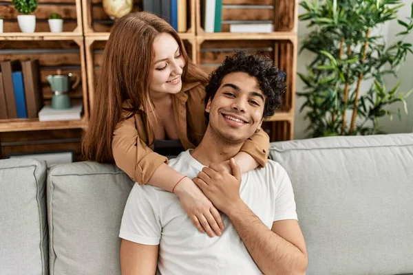 Mutlu gülümseyen ve sarılan genç çift evdeki kanepede oturuyor..