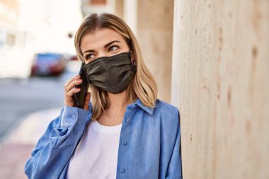Coronavirus maskesi takan genç sarışın kız şehirdeki akıllı telefondan konuşuyor..