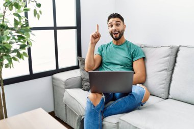Sakallı, bilgisayar bilgisayarı kullanan genç ve yakışıklı bir adam evdeki kanepede oturmuş başarılı bir fikirle işaret ediyor. Heyecanlı ve mutlu. Bir numara.. 