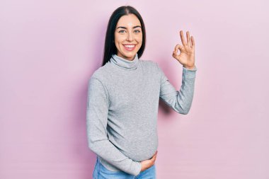 Mavi gözlü güzel bir kadın bebek bekliyor, hamile karnına dokunuyor gülümsüyor ve el ve parmaklarıyla tamam işareti yapıyor. başarılı ifade. 