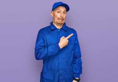 Sakallı, inşaatçı tulumu giymiş kel bir adam endişeli ve endişeli bir ifade takınıyor. 