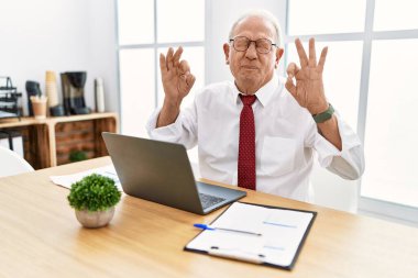 Ofiste çalışan kıdemli bir adam bilgisayarlı dizüstü bilgisayar kullanıyor gözleri kapalı gülümsüyor parmaklarıyla meditasyon hareketi yapıyor. yoga konsepti. 