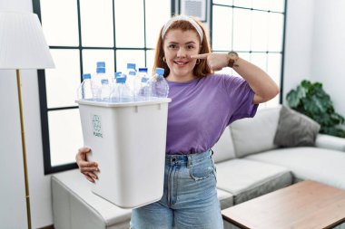Geri dönüşüm sepetini elinde tutan genç, kızıl saçlı bir kadın. Elinde plastik şişelerle, parmakları burnuna ve yüzüne dönük, neşeli bir şekilde gülümsüyor. güzellik konsepti 