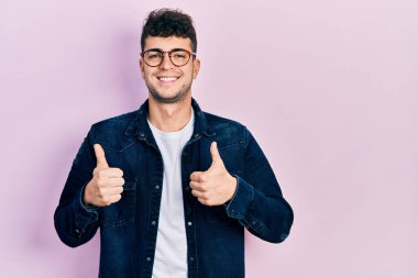 Gündelik kıyafetler giyen ve gözlük takan genç İspanyol adam. Başarıyla olumlu jest yapıyor. Gülümsüyor ve mutlu. Neşeli ifade ve kazanan jest. 