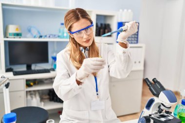 Genç sarışın kadın bilim adamı üniforması giyiyor. Laboratuvarda pipetle sıvı döküyor.