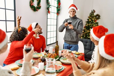 Mutlu bir şekilde gülümseyen bir grup genç Noel yemeği yiyor ve evde kadeh kaldırıyor..