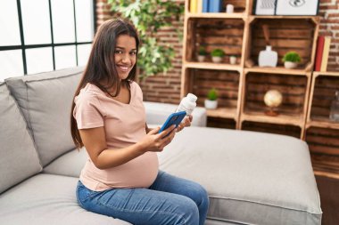Genç Latin kadın hamile. Evde elinde bir kutu hapla akıllı telefon kullanıyor.