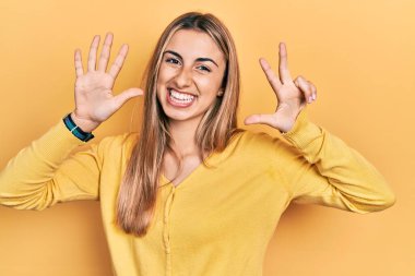 Güzel İspanyol kadın gündelik sarı süveter giyer ve 8 numaralı parmaklarıyla işaret ederken kendinden emin ve mutlu bir şekilde gülümser.. 