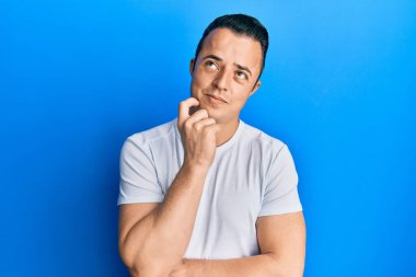 Yakışıklı, beyaz tişört giyen, şüpheye odaklanmış, çenesinde parmak olan ve kafasını kaldırıp merak eden genç bir adam. 