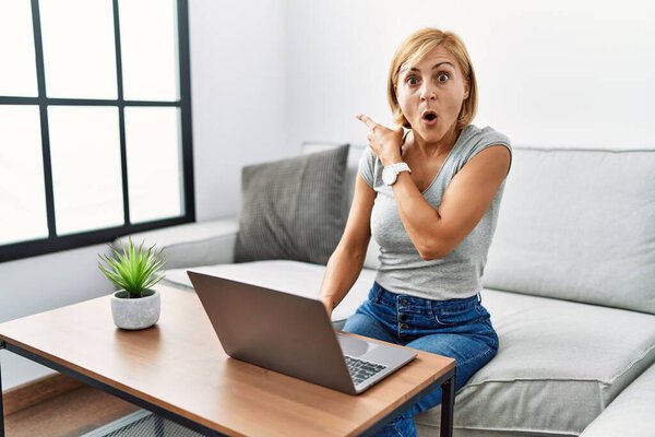 Блондинка средних лет с помощью ноутбука дома удивлен указывая пальцем в сторону, открытый рот удивлен выражение. 