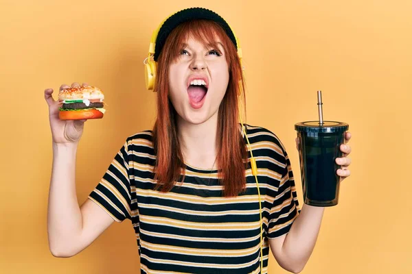 Redhead Young Woman Eating Burger Drinking Soda Wearing Headphones Angry — Stock Photo, Image